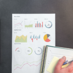 close up of spiral bound notebook and chart with graphs and someone's hand holding a pen, presumable working with the data