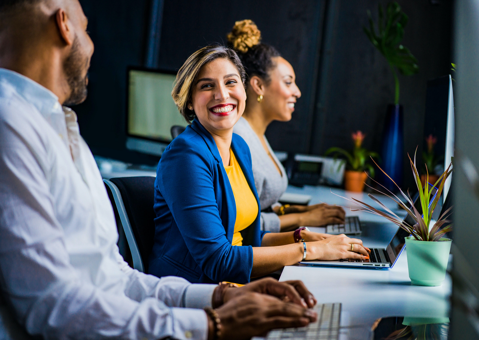 Woman smiling in centre, looking at her male colleage while another happy femail colleague is staring at her screen